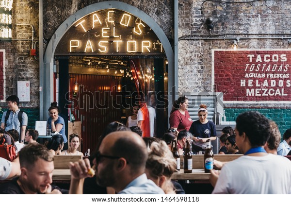 London,\
UK - July 23, 2019: People sitting at the outdoor tables of El\
Pastor Tacos restaurant in Borough Market, one of the largest and\
oldest food markets in London. Selective\
focus.