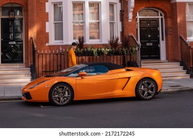 London, UK - June 11, 2022: Young man in orange shirt walks past an orange Lamborghini Gallardo Spyder, parked in Herbert Crescent area, London.