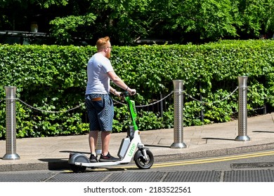 London, UK - June 2022: Person On An Electric Scooter Riding On A Road Past A Green Hedge Around Gardens In Canary Wharf