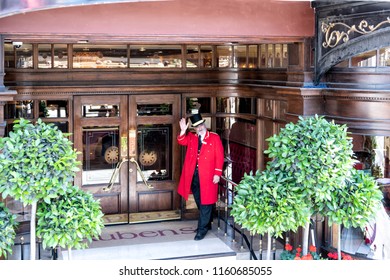 London, UK - June 22, 2018: Porter, Doorman In Traditional Attire, Clothing, Hat, Red Gown, Coat Standing At Rubens Hotel, Inn Entrance Waving Hand