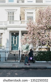 London, UK - March 27 2019: Mother And Little Daughter Riding The Scooter Walking On Sidewalk In Front Posh Residential Building. Family Lifestyle Concept. Spring In The City. City Street View 