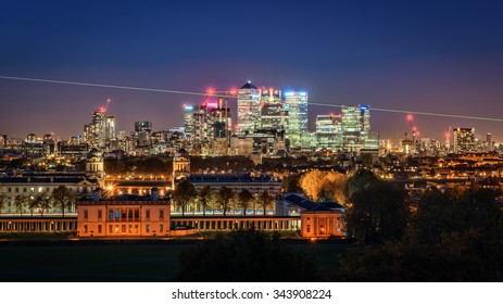 London, UK - October 25, 2015: High Visible Prime Meridian Beam At Night In Greenwich Park And With The Canary Wharf Skyline With Its Corporate Office Buildings In The Background.