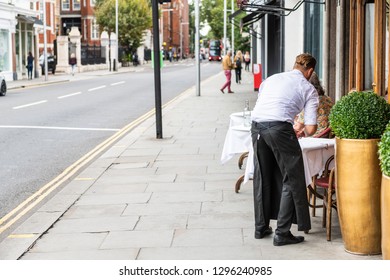 London, UK - September 16, 2018: Man Waiter Standing Setting Up Cafe Table In Chelsea Outside Restaurant Street Sidewalk During Day