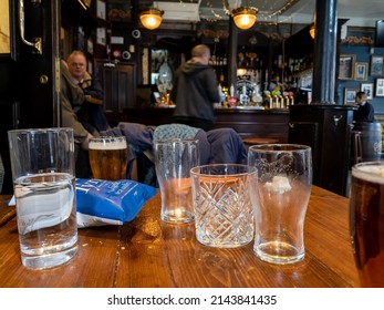 London. UK-04.05.2022. Empty And Full Pint Glasses Of Beer And Spirit And A Packet Of Crisp On A Table In An English Pub.