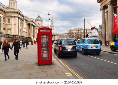 London, United Kingdom - March 6 2022: A Back London Taxi Cab Passes A Red British Phone Booth With People Passing And The National Gallery In The Background