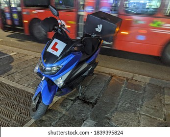 London/ United Kingdom - October 22 2020: Learner Delivery Motorbike/ Moped With Deliveroo Basket In Front Of A Moving London Bus At Night. Shows Gig Economy And Popularity Of Take Away Food. 