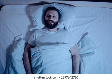 A lone bearded dark-haired man sleeps soundly lying on his back at night in bed. The general plan from above, white bedding, a bedroom, self-isolation.One - Shutterstock ID 1697642710