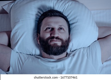 Lone bearded man cannot sleep in bed at night eyes wide open looking at the ceiling. Insomnia, pajamas, white bedding. Close up - Shutterstock ID 1677107461