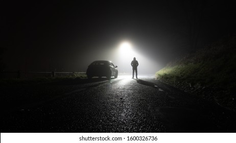 A Lone Car, Parked On The Side Of The Road, Underneath A Street Light, With A Hooded Figure, On A Spooky, Scary, Rural, Country Road. On A Foggy Winters Night