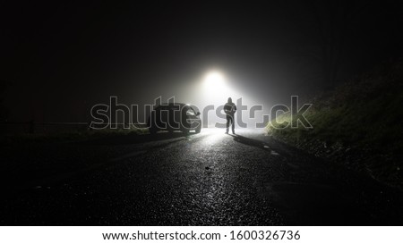 A lone car, parked on the side of the road, underneath a street light, with a hooded figure, on a spooky, scary, rural, country road. On a foggy winters night