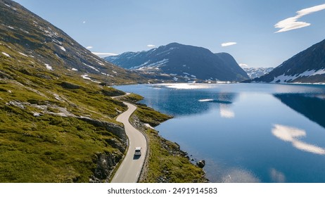 A lone car winds its way along a mountain road overlooking a shimmering blue lake, with snow-capped peaks rising in the distance. Langvatnet, Geiranger, Norway - Powered by Shutterstock