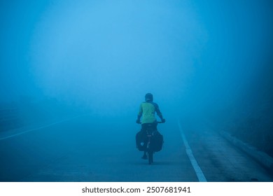 A lone cyclist navigates through a thick, blue mist on a desolate road, creating a surreal and atmospheric scene that embodies solitude and endurance. - Powered by Shutterstock