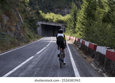 A lone cyclist rides on a winding road through a mountain pass. The road is surrounded by trees and rocks, creating a sense of isolation and adventure. Cycling adventure in Romania. - Powered by Shutterstock