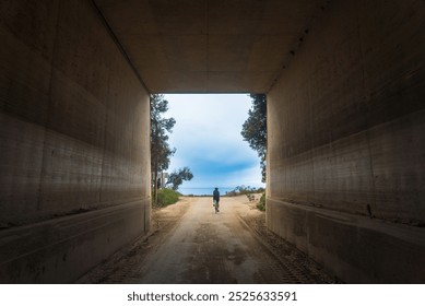 A lone cyclist rides through a concrete tunnel towards an open sky, framed by trees and a road ahead, symbolizing journey, adventure, and exploration. - Powered by Shutterstock