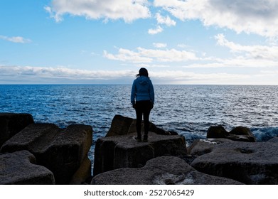 A lone female figure stands on large artificial coastal rocks, gazing out over the vast atlantic ocean beneath a bright blue sky. - Powered by Shutterstock