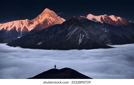 A lone figure stands atop a hill, surrounded by clouds, facing a dramatic mountain range with a striking, sunlit peak. - Powered by Shutterstock