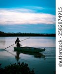 A lone figure stands on a boat, skillfully maneuvering a pole against the tranquil waters of the lake. The soft hues of dawn reflect on the surface, creating a serene atmosphere