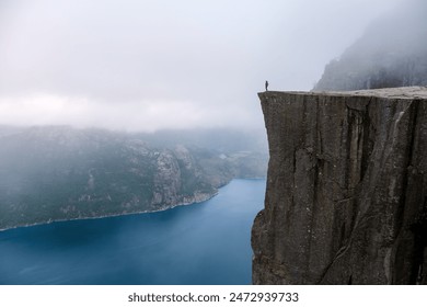 A lone figure stands on the edge of Preikestolen, a dramatic cliff in Norway, overlooking a vast expanse of water shrouded in mist. Preikestolen, Norway - Powered by Shutterstock