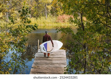 A lone figure walks down a wooden dock with a colorful surfboard, amidst vibrant autumn foliage by a serene lake, ideal for adventure seekers and nature lovers - Powered by Shutterstock