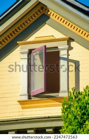 A lone hidden windows on attic bedroom on top story of house in late afternoon sun with front yard trees and blue sky background. Open glass on hindge with yellow wooden home exterior neighborhood.