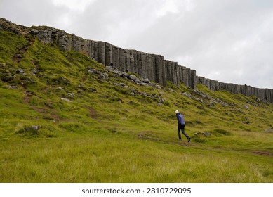 Lone hiker in a red jacket walking beside towering basalt columns and cliffs in a lush green rift valley