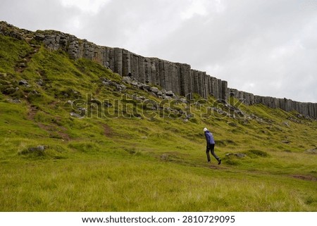 Lone hiker in a red jacket walking beside towering basalt columns and cliffs in a lush green rift valley
