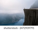 A lone hiker stands on the edge of Preikestolen, a dramatic cliff overlooking a misty fjord in Norway. Preikestolen Norway