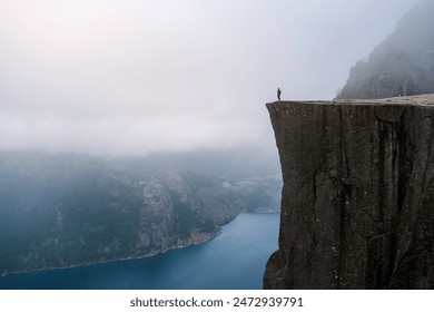 A lone hiker stands on the edge of Preikestolen, a dramatic cliff overlooking a misty fjord in Norway. Preikestolen Norway - Powered by Shutterstock