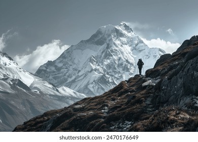 A lone hiker stands on a rugged trail, gazing at a majestic, snow-covered mountain peak under a cloudy sky. - Powered by Shutterstock