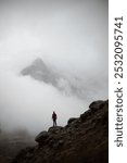 A lone hiker stands triumphantly on a rugged mountain peak, surrounded by fog and clouds.