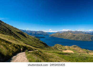 A lone hiker walks along a mountain path overlooking a vast, tranquil lake surrounded by rolling hills and distant mountains. The clear blue sky adds to the serene and expansive feeling of the landsc - Powered by Shutterstock
