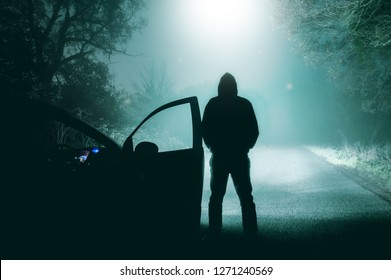 A Lone, Hooded Figure Standing Next To A Car Looking At A Light Beam Coming Down From A UFO On A Misty Country Road Silhouetted At Night By Car Headlights