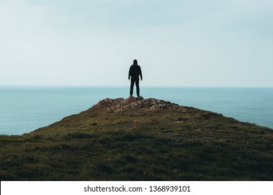 A lone hooded figure standing on top of a hill, looking out across the ocean.
