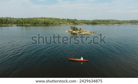 A lone kayaker in a red vessel glides through the water near a small, tree-covered island, the scene an embodiment of peaceful exploration and the beauty of seclusion in nature. Aerial view, kayak