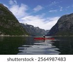 A lone kayaker in a vibrant red kayak paddling through the calm waters of Eidfjord in Norway, surrounded by steep, lush green cliffs and towering snow-capped mountains under a bright blue sky