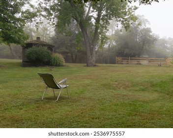 A lone lawnchair sits on the grassy lawn in a large backyard on a gloomy, foggy day. A gazebo and rach style fence are in the background along with trees.  - Powered by Shutterstock