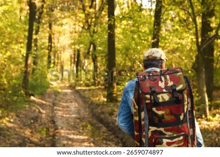 A lone man with a backpack is captured in a blurred background shot, hiking through a vibrant forest. This image evokes the spirit of summer or autumn travel and adventure.