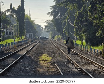 A lone man carrying a large sack walks along railway tracks under the morning sun, with lush greenery and a rural setting in the background. - Powered by Shutterstock