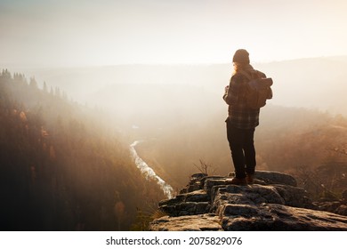A lone man in a flannel shirt with a backpack on his back stands on a rocky outlook in an autumn landscape during a misty sunset.