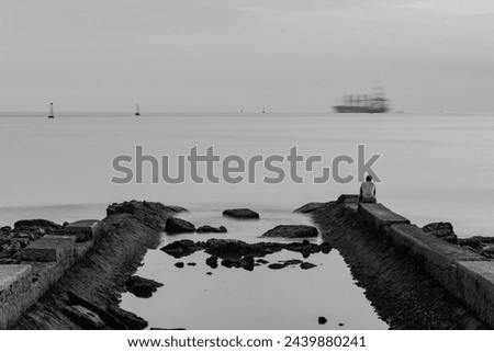 Lone man sitting on the wall of the water canal watching the sea. Late afternoon time lapse on the beach. Cargo ship passing on the horizon. Santos city, Brazil. Black and white image. 