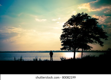 A lone man standing at the shore, beside a tree,  under a dramatic evening sky - Powered by Shutterstock