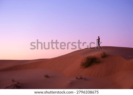 Lone man using mobile phone on top of a sand dune during a pastel purple sunset in the desert