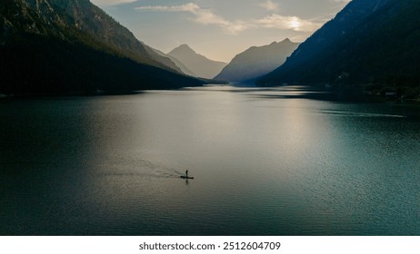 A lone paddleboarder glides over Plansee's calm waters at sunrise, as the sun casts a warm glow on the lake and highlights the stunning mountains surrounding this serene oasis. - Powered by Shutterstock