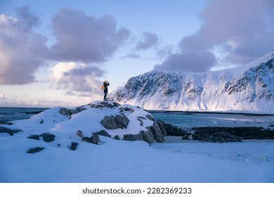 Lone person looks onwards at a mountain in snow during sunset with the peak visible. Scenic landscape photo composite. The stony beach and cliff of rocky bay. Blue tones of February sunset, 