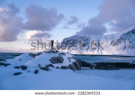 Lone person looks onwards at a mountain in snow during sunset with the peak visible. Scenic landscape photo composite. The stony beach and cliff of rocky bay. Blue tones of February sunset, 