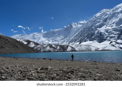 A lone person standing by a serene lake with a backdrop of snow-covered mountains under a clear blue sky. - Powered by Shutterstock