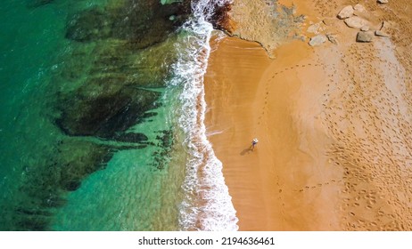 Lone Person Walking Along A Red Sand Beach From High Above In Contrasting Against The Clear Greenish Blue Waters Leaving Footprints