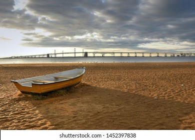 A Lone Rowboat Sets On An Empty Beach In The Early Morning Light.  The Small, Dry Docked Boat Points Toward The Chesapeake Bay Bridge On The Horizon.