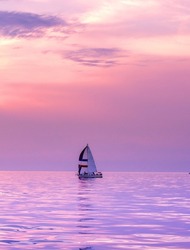 A Lone Sail Boat Floats On A Still Lake With A Beautiful Sunset Of Purple And Pink