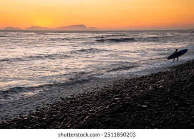 A lone surfer carrying a surfboard walks along a rocky beach as the sun sets, casting a beautiful golden glow and soft waves. - Powered by Shutterstock